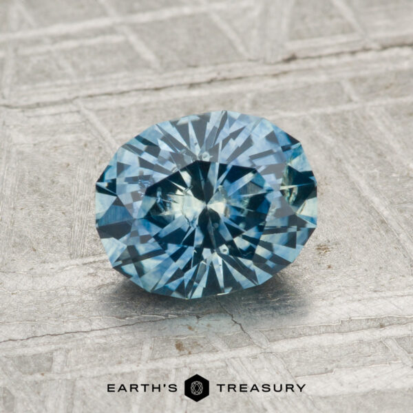 1.39-Carat Montana Sapphire (Heated) A 1.39-carat heated Montana sapphire, round brilliant-cut and blue, is shown on a textured gray surface with the "Earth's Treasury" logo at the bottom.