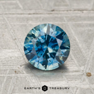 A 1.37-carat heated Montana sapphire with a round brilliant cut and blue-green color rests on a textured gray surface, with "EARTH'S TREASURY" shown below.