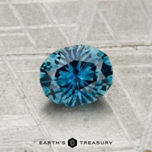 A 0.97-carat heated Montana Sapphire, brilliant-cut in teal, displayed on a metallic textured surface with "Earth's Treasury" beneath.