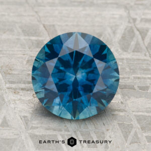 A 2.67-carat heated Montana sapphire, round brilliant-cut and blue, displayed on a textured grey surface with the Earth’s Treasury logo and text below.