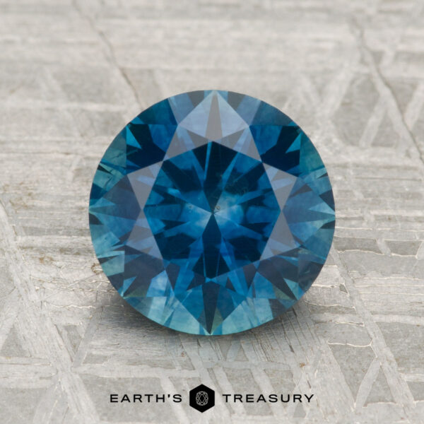 2.67-Carat Montana Sapphire (Heated) A 2.67-carat heated Montana sapphire, round brilliant-cut and blue, displayed on a textured grey surface with the Earth’s Treasury logo and text below.