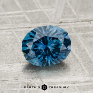 A round, faceted 1.39-carat Montana Sapphire (Heated) with a blue hue rests on a textured gray surface. The "Earth's Treasury" logo and text are displayed at the bottom of the image.