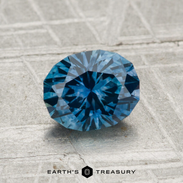 1.39-Carat Montana Sapphire (Heated) A round, faceted 1.39-carat Montana Sapphire (Heated) with a blue hue rests on a textured gray surface. The "Earth's Treasury" logo and text are displayed at the bottom of the image.