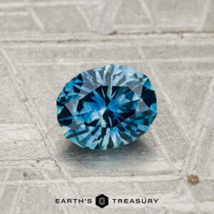 A 0.94-carat heated Montana sapphire, faceted in an oval shape, rests on a textured metallic surface with "Earth's Treasury" and a geometric logo below.