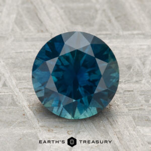 A 2.52-carat heated Montana sapphire, round-cut and blue, is displayed on a textured gray surface with "EARTH'S TREASURY" at the bottom.