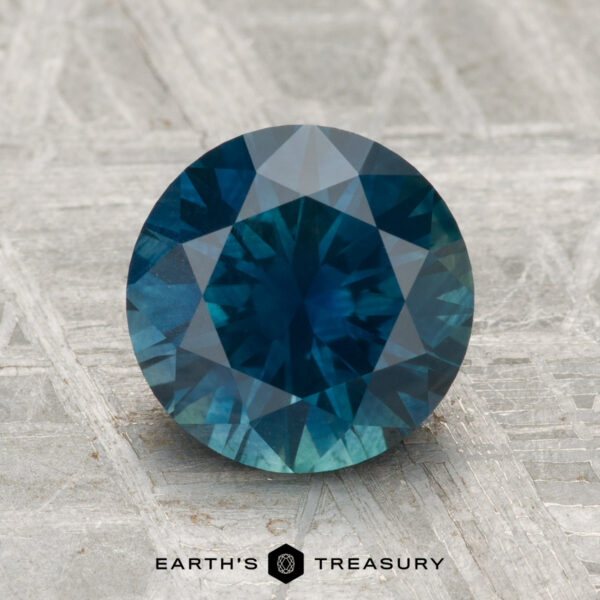 2.52-Carat Montana Sapphire (Heated) A 2.52-carat heated Montana sapphire, round-cut and blue, is displayed on a textured gray surface with "EARTH'S TREASURY" at the bottom.