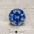 A 1.36-carat heated Montana sapphire, round brilliant-cut, is displayed on a textured light surface with “Earth's Treasury” branding at the bottom.