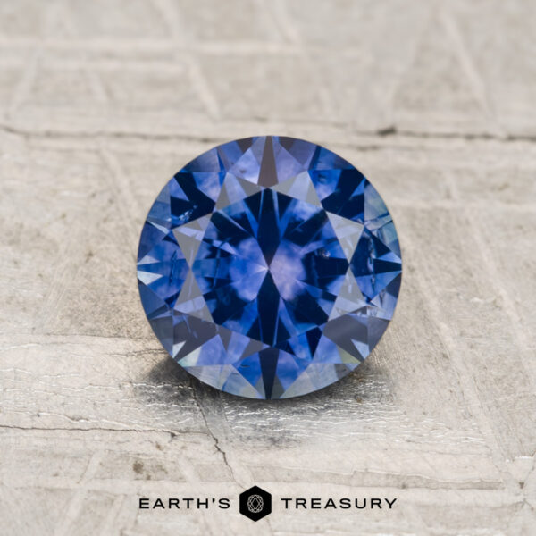 1.36-Carat Montana Sapphire (Heated) A 1.36-carat heated Montana sapphire, round brilliant-cut, is displayed on a textured light surface with “Earth's Treasury” branding at the bottom.