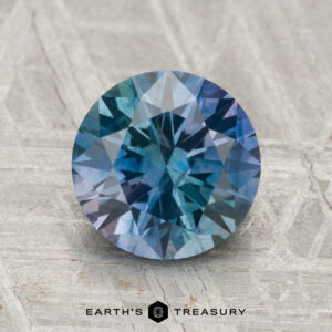 A 2.44-carat heated Montana Sapphire, cut in a round brilliant style with blue-green hues, displayed upright on a textured gray surface above the Earth's Treasury logo.