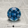 1.36-carat heated Montana sapphire in a round brilliant cut displayed on a textured gray surface, with "Earth's Treasury" at the bottom.