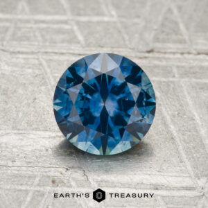 1.36-carat heated Montana sapphire in a round brilliant cut displayed on a textured gray surface, with "Earth's Treasury" at the bottom.