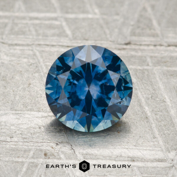 1.36-Carat Montana Sapphire (Heated) 1.36-carat heated Montana sapphire in a round brilliant cut displayed on a textured gray surface, with "Earth's Treasury" at the bottom.