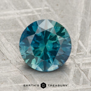 A 2.42-carat heated Montana Sapphire, cut in a round brilliant shape with blue-green hues, is displayed on a textured gray surface featuring the text "EARTH'S TREASURY" below.