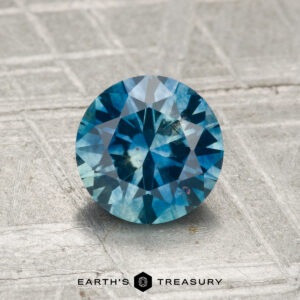 A 1.35-carat heated Montana sapphire in round brilliant cut is displayed on a textured gray surface, with "EARTH'S TREASURY" shown at the bottom.