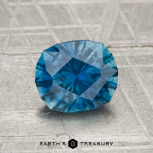 A 1.66-carat heated Montana sapphire with a faceted blue surface rests on textured gray, accompanied by "EARTH'S TREASURY" text and hexagon logos below.