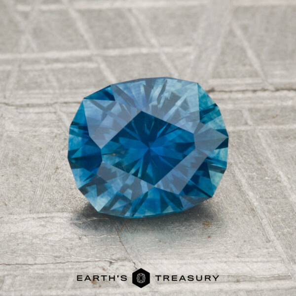 1.66-Carat Montana Sapphire (Heated) A 1.66-carat heated Montana sapphire with a faceted blue surface rests on textured gray, accompanied by "EARTH'S TREASURY" text and hexagon logos below.