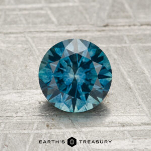 A 1.34-carat heated Montana sapphire, round brilliant-cut and blue, is shown on a textured metallic surface with "EARTH'S TREASURY" displayed below.