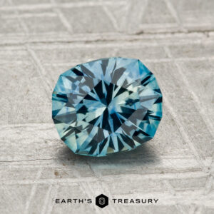 A 1.39-carat heated Montana sapphire, round and blue-green, is displayed on a textured metallic surface with the Earth’s Treasury logo below.