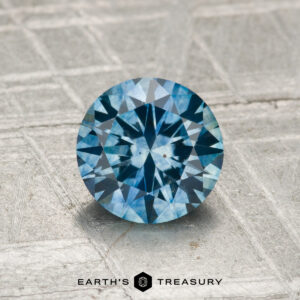 A 1.31-carat heated Montana sapphire, round brilliant-cut and blue, showcased on a textured metallic surface with "Earth's Treasury" displayed at the bottom.
