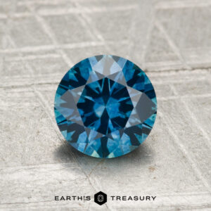 A 1.31-carat heated Montana sapphire with a round brilliant cut is displayed on a textured grey surface, with "Earth's Treasury" visible at the bottom.