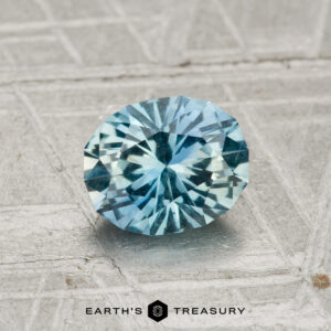 A 1.19-Carat Montana Sapphire gemstone is shown on a metallic, textured surface with "EARTH'S TREASURY" displayed at the bottom.
