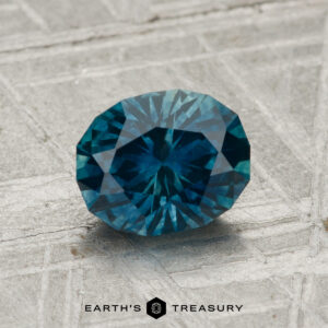 A 1.33-carat heated Montana sapphire is showcased on a textured gray surface with "EARTH'S TREASURY" and a logo at the bottom.