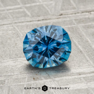 A 1.37-carat heated Montana sapphire with an oval cut rests on a textured gray surface; the "Earth's Treasury" logo appears at the bottom.
