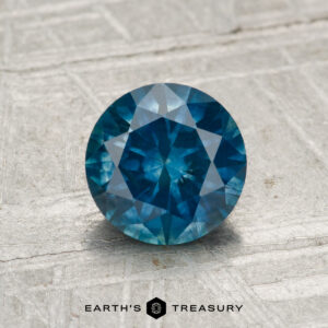 A round, faceted 1.31-carat heated Montana sapphire is displayed on a textured light gray surface with the text "EARTH'S TREASURY" below it.