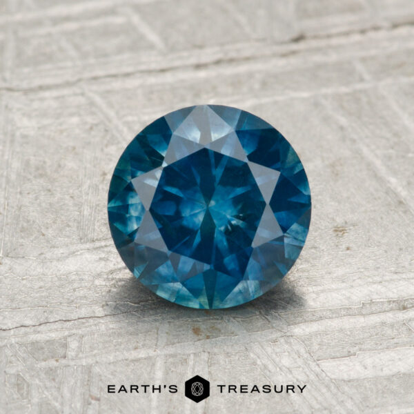 1.31-Carat Montana Sapphire (Heated) A round, faceted 1.31-carat heated Montana sapphire is displayed on a textured light gray surface with the text "EARTH'S TREASURY" below it.