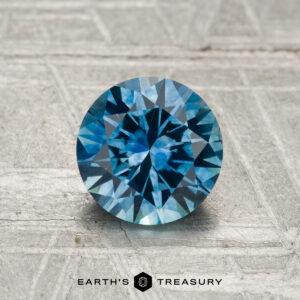 A 1.29-carat heated Montana sapphire, cut in a round brilliant shape, is shown on a textured gray surface with “Earth’s Treasury” at the bottom.