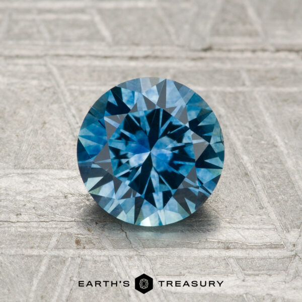 1.29-Carat Montana Sapphire (Heated) A 1.29-carat heated Montana sapphire, cut in a round brilliant shape, is shown on a textured gray surface with “Earth’s Treasury” at the bottom.