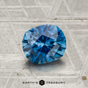 A 1.36-carat heated Montana Sapphire rests on a textured metallic surface, with "Earth's Treasury" and the logo visible at the bottom.