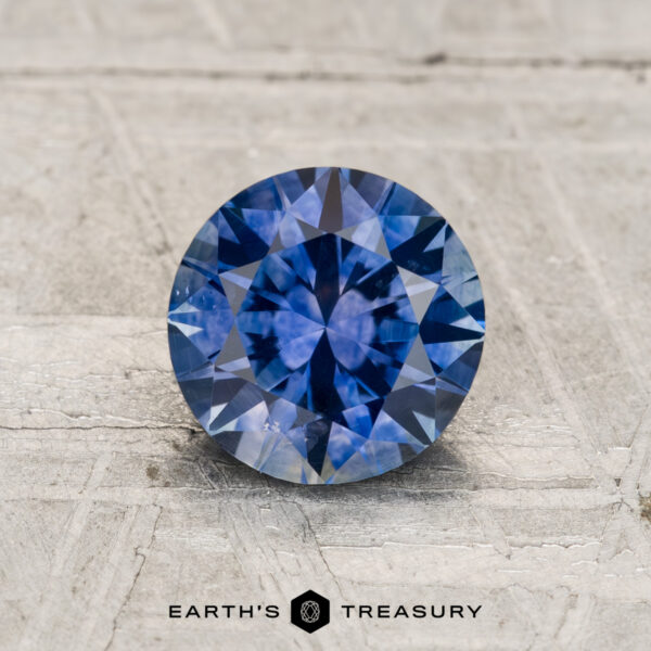 1.29-Carat Montana Sapphire (Heated) A round, faceted 1.29-carat heated Montana sapphire with a blue hue sits on a textured, light surface featuring the "EARTH'S TREASURY" logo and text at the bottom.
