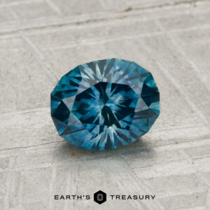 A 1.31-carat, oval-shaped, blue-green faceted Montana sapphire (heated) rests on a light textured surface, with “Earth’s Treasury” displayed at the bottom.