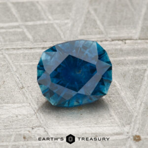 A 1.32-carat heated Montana sapphire is shown on a textured gray surface with "EARTH'S TREASURY" at the bottom.