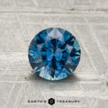 A 1.29-carat heated Montana sapphire, round brilliant-cut and blue, is displayed on a textured surface with the text "EARTH'S TREASURY" below.