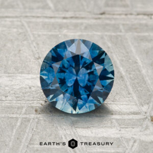 A 1.29-carat heated Montana sapphire, round brilliant-cut and blue, is displayed on a textured surface with the text "EARTH'S TREASURY" below.