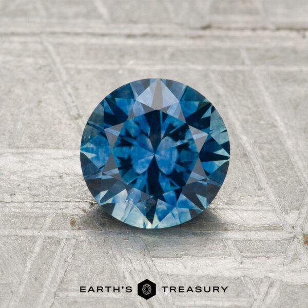 1.29-Carat Montana Sapphire (Heated) A 1.29-carat heated Montana sapphire, round brilliant-cut and blue, is displayed on a textured surface with the text "EARTH'S TREASURY" below.