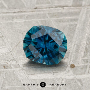 A 1.23-carat heated Montana sapphire rests on a textured metallic surface with the text "EARTH'S TREASURY" and a hexagonal logo below.
