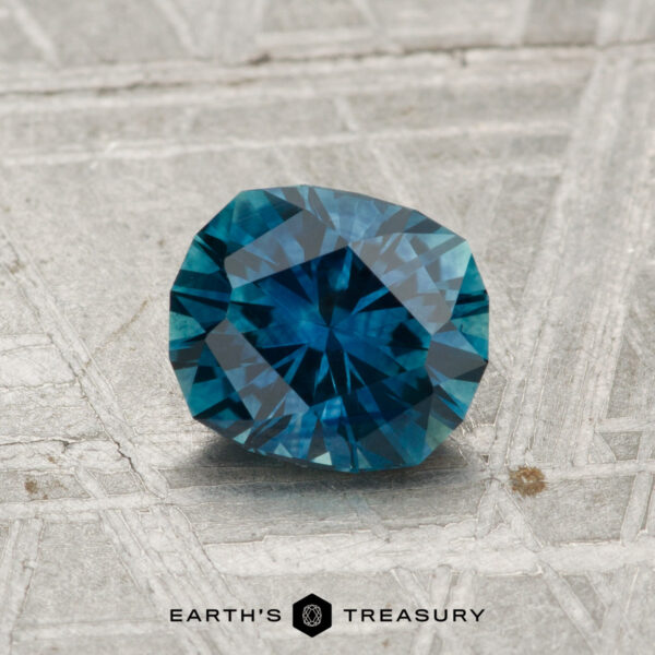 1.23-Carat Montana Sapphire (Heated) A 1.23-carat heated Montana sapphire rests on a textured metallic surface with the text "EARTH'S TREASURY" and a hexagonal logo below.