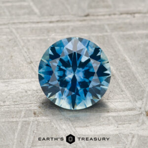A 1.28-carat heated Montana Sapphire (round brilliant-cut, blue) rests on a textured gray surface with "EARTH'S TREASURY" at the bottom.