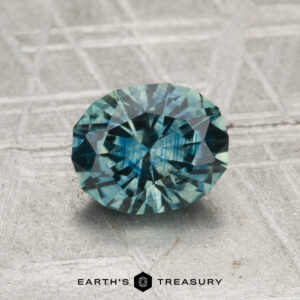 A 1.31-carat Montana sapphire (heated), oval-cut and blue-green, displayed on a textured gray surface with "EARTH'S TREASURY" at the bottom.