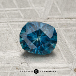 A 1.22-carat heated Montana sapphire with a faceted blue gem sits on a textured gray surface, featuring the "Earth's Treasury" text at the bottom.