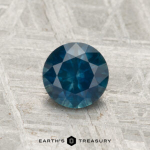 A 0.92-carat round-cut heated Montana sapphire with multiple facets is displayed on a textured light gray surface. "EARTH'S TREASURY" appears at the bottom.