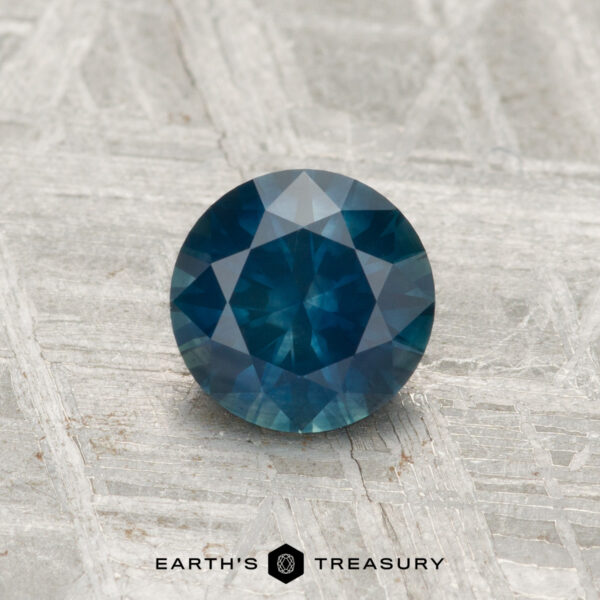 0.92-Carat Montana Sapphire (Heated) A 0.92-carat round-cut heated Montana sapphire with multiple facets is displayed on a textured light gray surface. "EARTH'S TREASURY" appears at the bottom.