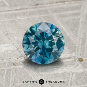 A 1.28-carat heated Montana sapphire with a round cut and blue-green hue rests on a textured gray surface, featuring the “Earth’s Treasury” label at the bottom.