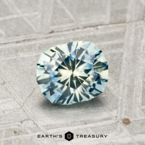A 1.20-carat heated Montana sapphire with blue-green hues, cut in a faceted oval shape, rests on a textured metallic surface. The "EARTH'S TREASURY" text and logo appear at the bottom.
