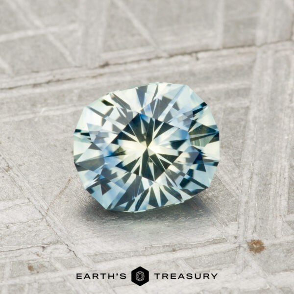 1.20-Carat Montana Sapphire (Heated) A 1.20-carat heated Montana sapphire with blue-green hues, cut in a faceted oval shape, rests on a textured metallic surface. The "EARTH'S TREASURY" text and logo appear at the bottom.