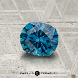 A 1.20-carat heated Montana sapphire is showcased on a textured gray surface with "EARTH'S TREASURY" at the bottom.