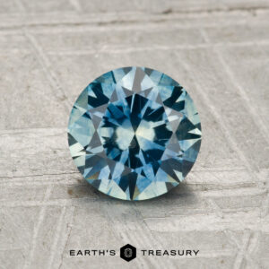 A 1.27-carat heated Montana Sapphire, round brilliant-cut and blue, is displayed on a textured gray surface with "Earth's Treasury" at the bottom.
